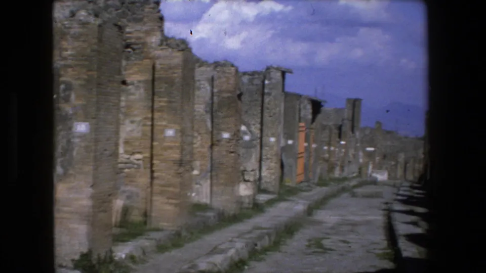 1973 8mm footage in Italy showing a narrow stone pathway between weathered brick and stone ruins beneath a bright blue sky.