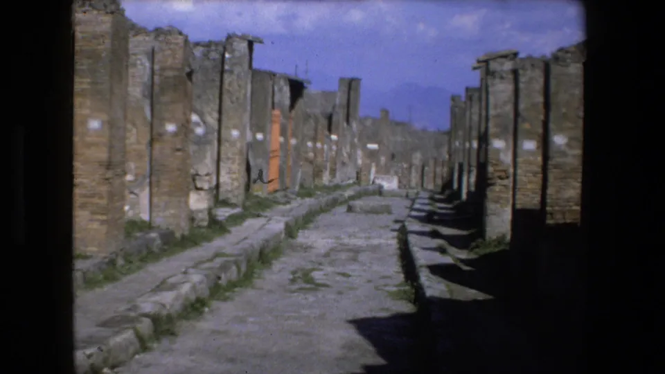 Handheld 8mm view of an ancient stone path bordered by tall weathered ruins and deep side shadows in Italy.