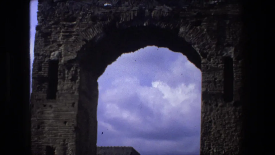 A large ancient masonry arch in shadow frames blue sky and clouds, continuing the 1973 Italy ruins sequence.