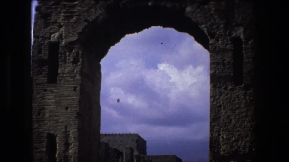 1973 Rome 8mm frame showing a shadowed weathered stone arch with blue cloudy sky and low ruin walls beyond it.