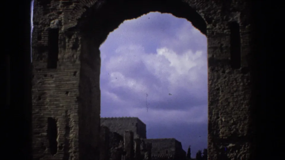1973 Rome travel footage frame with a dark arched ruin opening, textured masonry sides, and distant stone structures under a clouded sky.