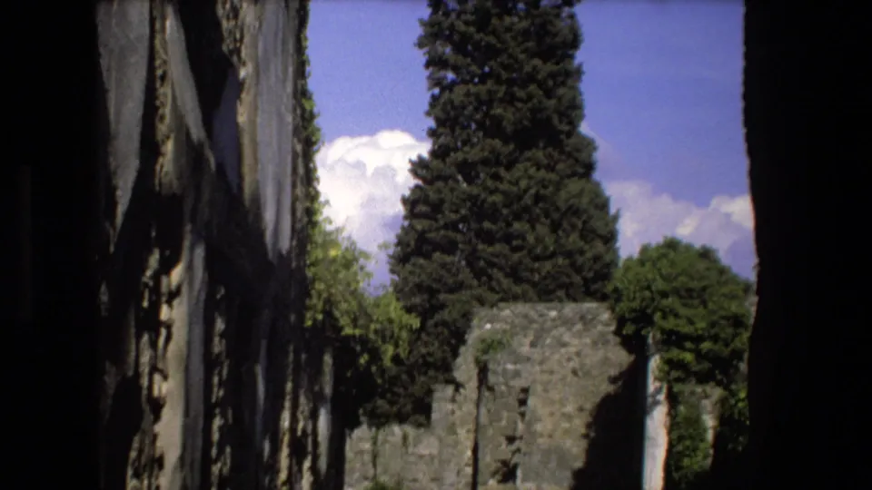 1973 Rome home-movie frame with a tall evergreen, crumbling stone wall, surrounding greenery, and bright clouds above the ruins.