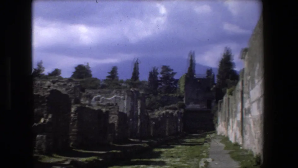 1973 Rome home-movie frame showing ruined masonry along a path, with cypress-like trees and heavy cloud cover in the background.
