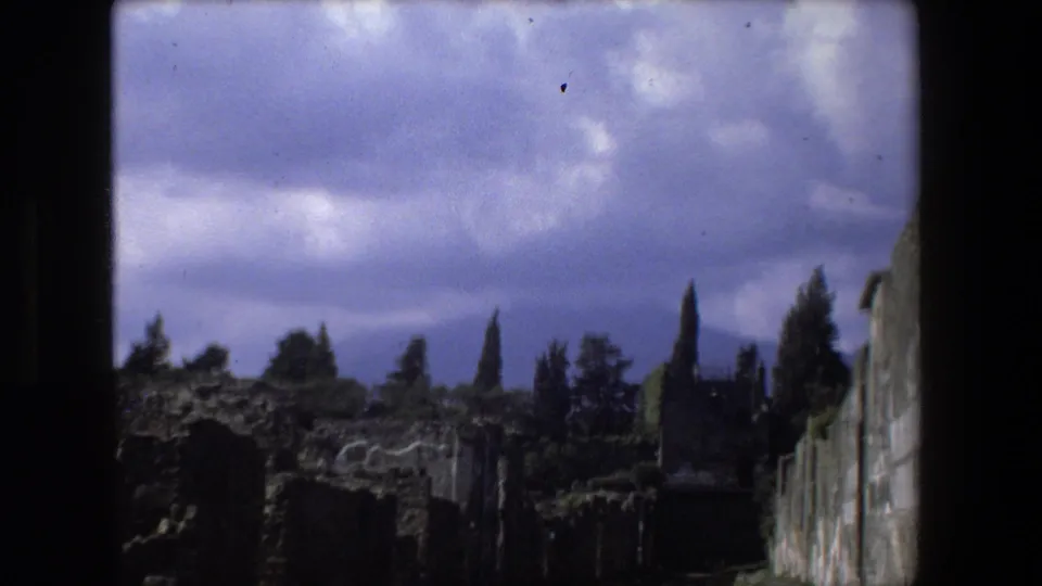 1973 Rome frame with underexposed home-movie texture, showing ancient stone ruins, trees, grass, and a dramatic cloudy sky.