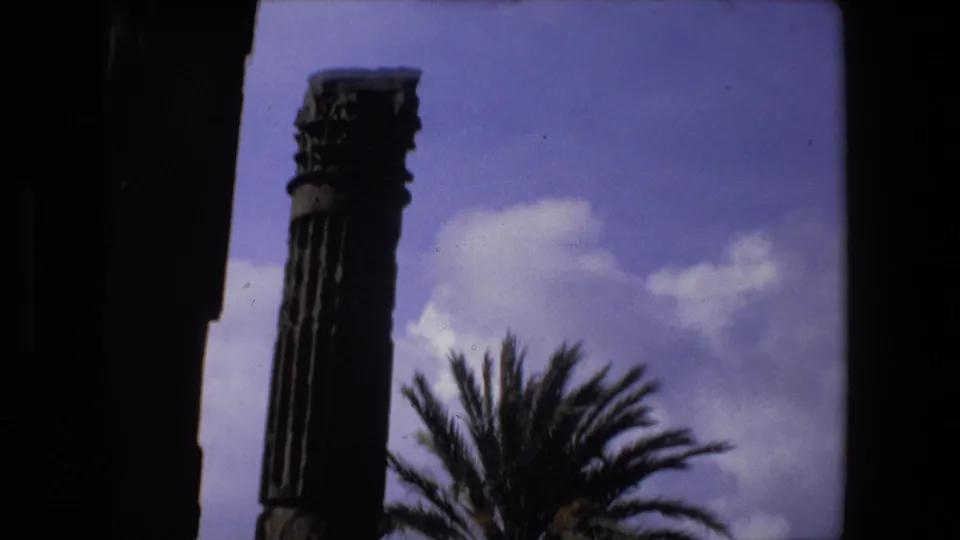 1973 Rome 8mm frame with a tall ancient column, palm fronds, partial ruin walls, and a blue-purple sky with clouds.