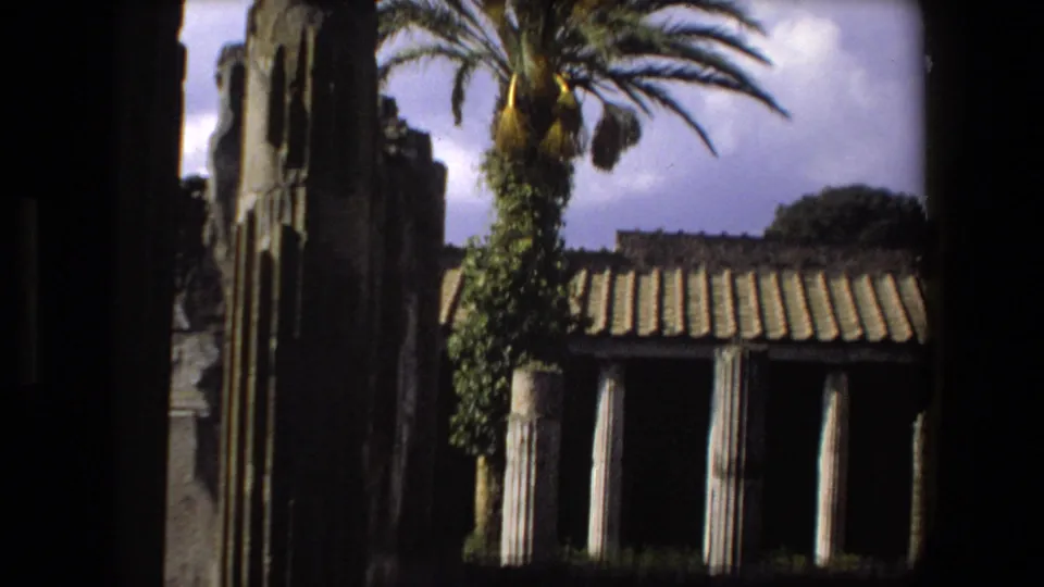 1973 Rome 8mm frame showing a palm tree beside ancient stonework, a column edge, and a small tiled-roof ruin structure under clouds.