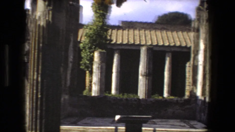 1973 Rome home-movie frame looking from shadow toward ancient stone columns, a tiled roof, greenery, and a paved courtyard area.