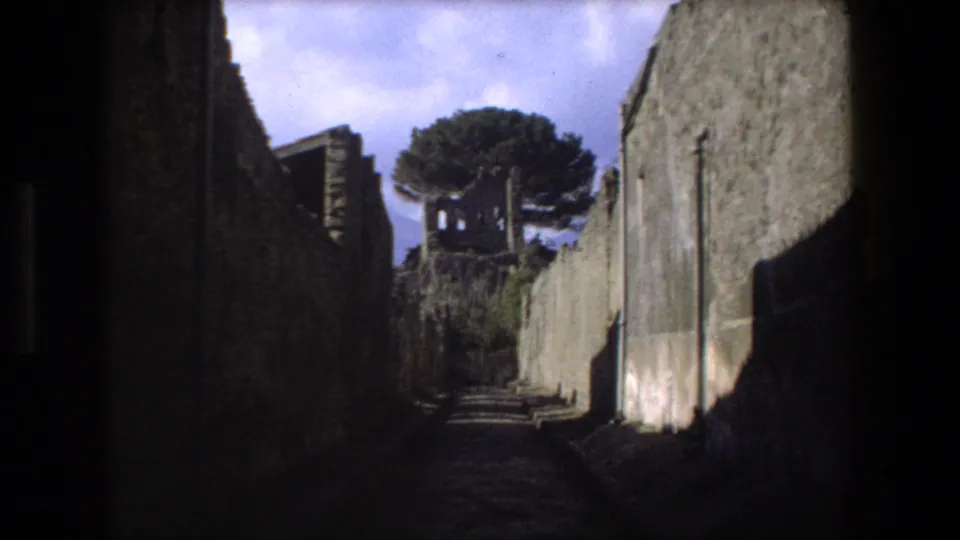 1973 Rome 8mm frame of a darkened stone path flanked by high ruin walls, leading toward a tree and weathered architectural remains.