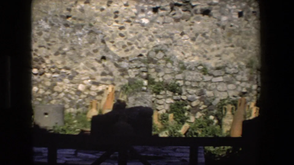 Close 1973 Rome home-movie view of weathered stone masonry, low greenery, and scattered architectural fragments near the base of the wall.