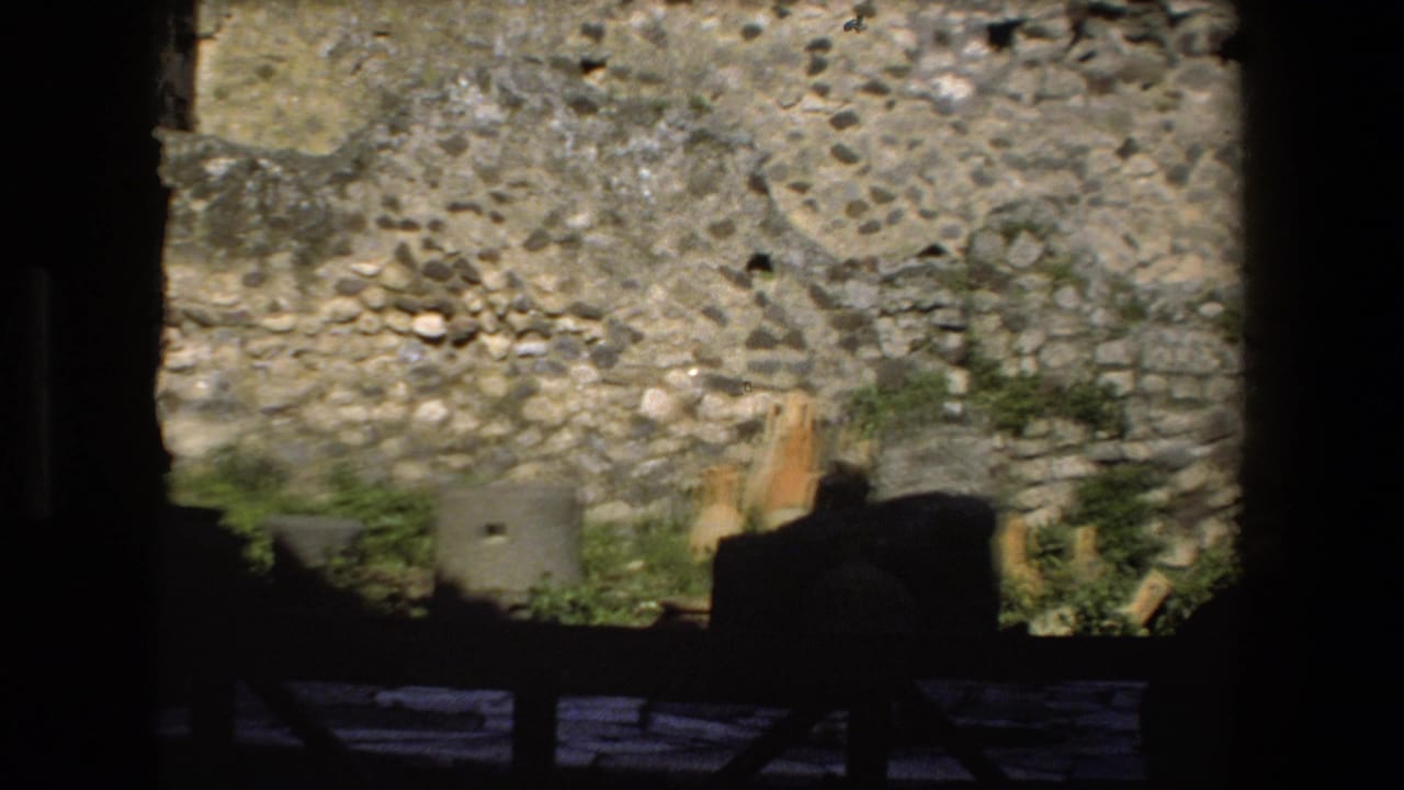 A handheld 8mm view in Rome, Italy, shows a sunlit rough stone wall seen through a dark frame, with shadowed ruin structures in the foreground.