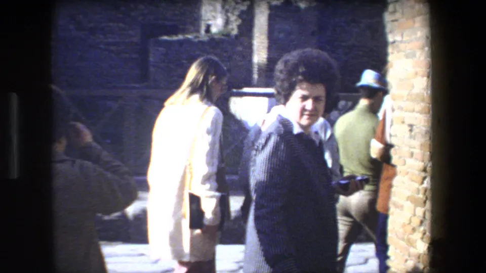 In Rome, Italy, tourists walk through a sunlit ruin area, with a woman in a dark patterned jacket and voluminous hairstyle looking toward the camera.