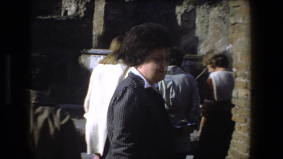 A 1973 Rome home-movie frame shows tourists beside brick and stone ruins, including a woman in profile near the camera holding a small dark object.