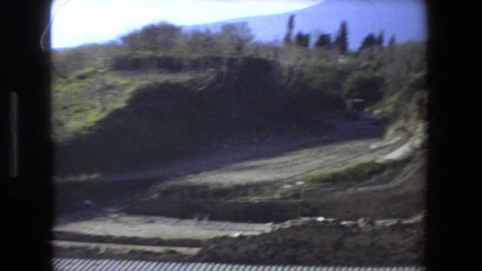 Handheld 1973 Rome home-movie frame showing a blurred dirt path, earth embankment, low stone edges, and trees around the ruin site.
