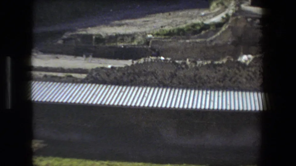 1973 Rome frame looking past a dark foreground edge and corrugated surface toward earth, paths, low walls, and vegetation within the ruin grounds.