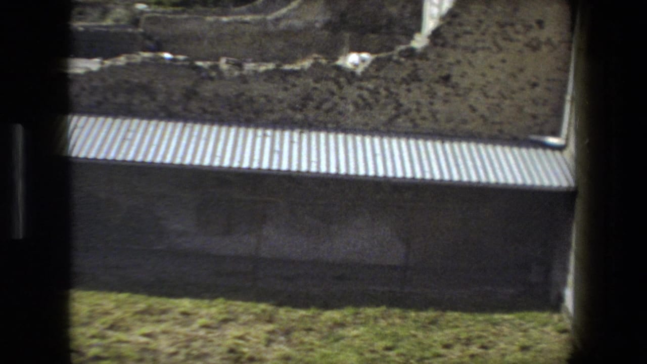Rome, Italy 1973 home-movie frame showing a corrugated protective roof below exposed earth and grass at a historic excavation site.