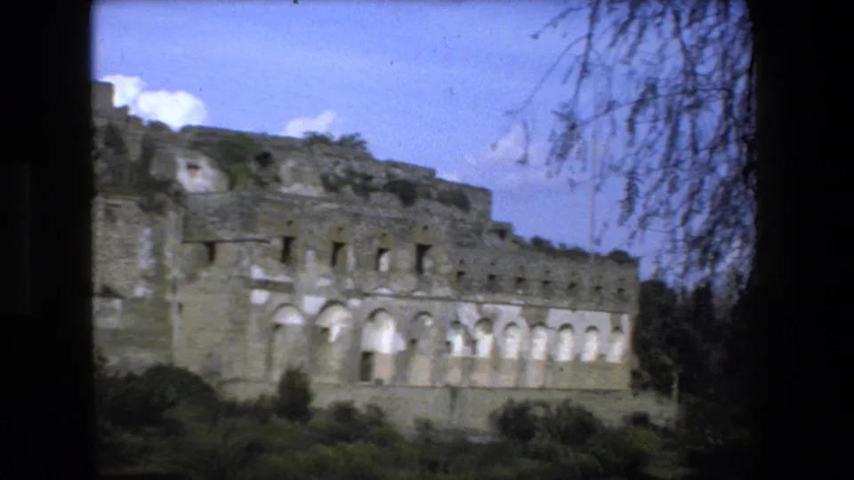 Rome, Italy 1973 frame showing a partially unearthed stone structure with arched openings, vegetation, blue sky, and film-gate edges.
