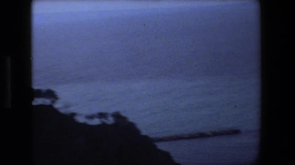 1973 Rome-area 8mm frame looking past a dark rocky shoreline or cliff edge toward a calm blue body of water, likely filmed as part of the same travel sequence.