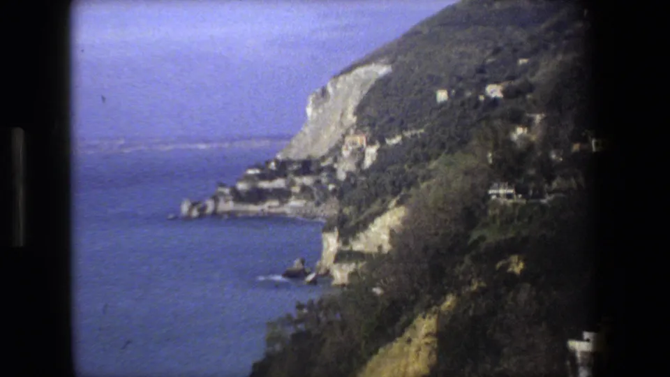 A clearer 1973 8mm frame shows a steep Mediterranean coastal hillside, pale cliffs, scattered shoreline buildings, and blue water viewed from a moving automobile.