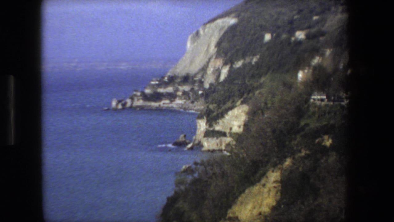 1973 8mm home-movie view of an Italian coastal bluff with deep blue water, wooded slopes, pale rock faces, and a road or structures along the hillside.