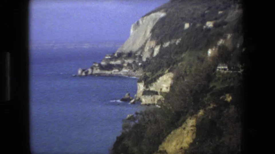 1973 8mm home-movie view of an Italian coastal bluff with deep blue water, wooded slopes, pale rock faces, and a road or structures along the hillside.