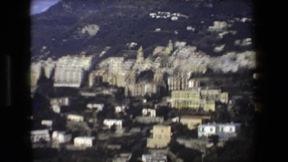 1973 home-movie frame showing a dense Italian hillside urban area with pale apartment blocks or hotels set below dark wooded slopes near the coast.