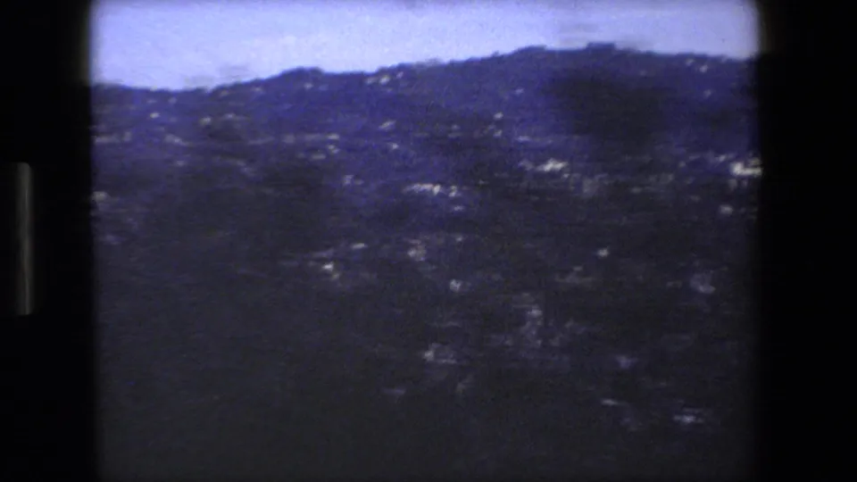 Soft 8mm view across a shadowed Italian hillside with scattered light buildings, tree cover, and a distant ridge under a pale sky.