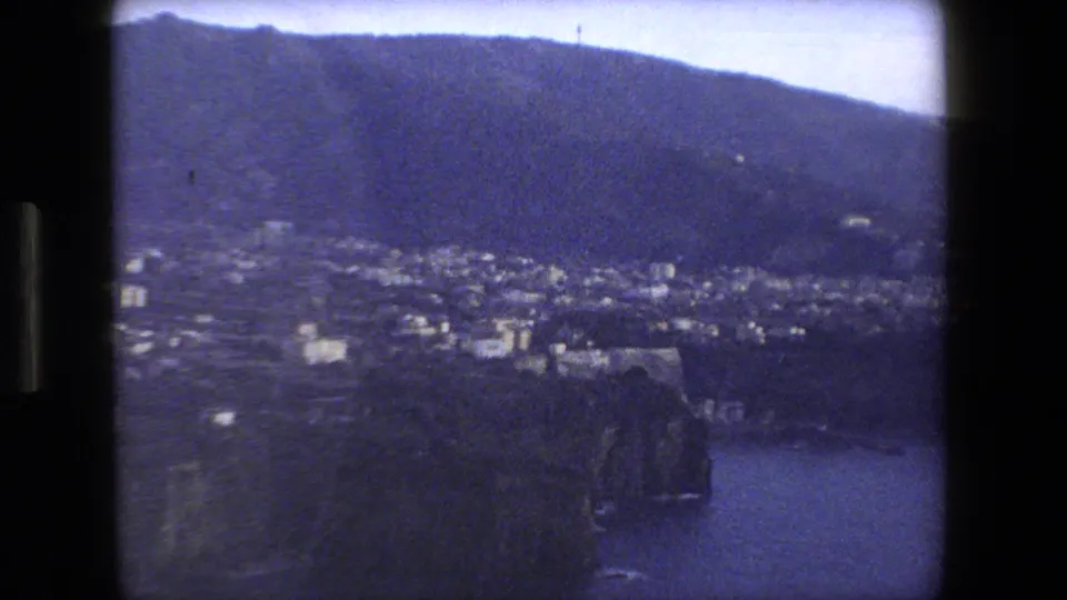 Wide 1973 8mm travel view of an Italian coastal settlement spread across a hillside, with a broad mountain slope behind it and water visible at the lower right.
