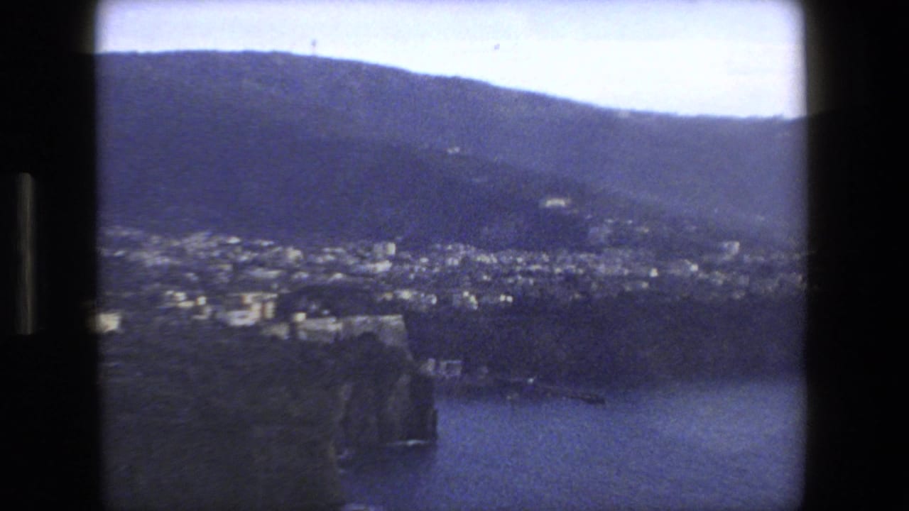 1973 8mm home-movie frame showing a Rome, Italy coastal area with clustered buildings near steep cliffs, dark hills, and water below.
