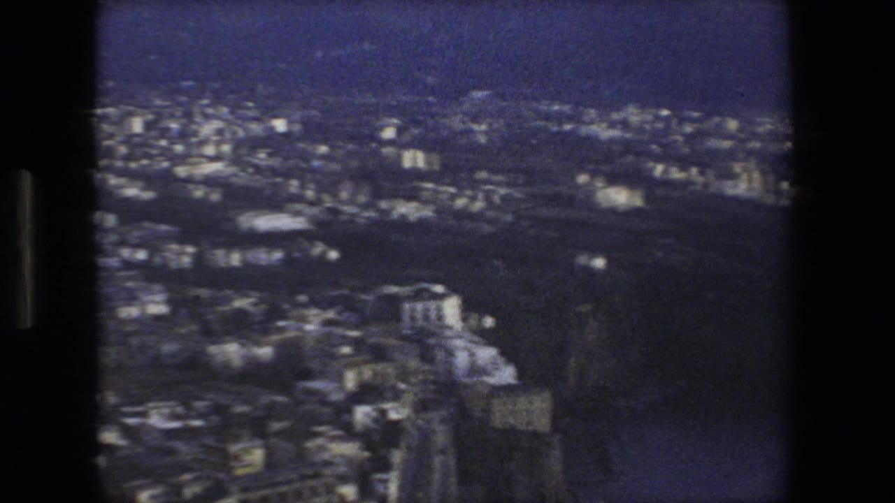 Soft 8mm frame of a Rome-area Italian hillside cityscape with dense buildings, roads, and dark terrain beyond.