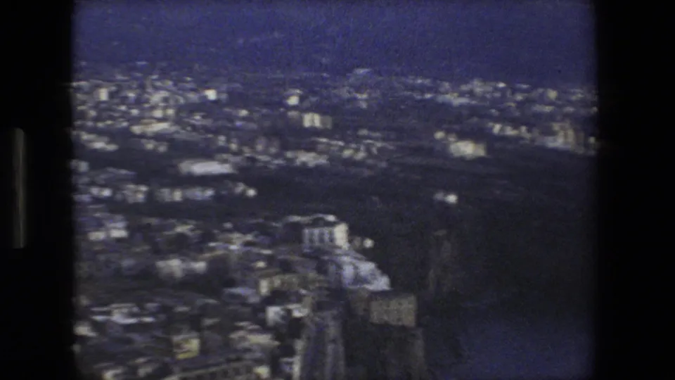 Soft 8mm frame of a Rome-area Italian hillside cityscape with dense buildings, roads, and dark terrain beyond.