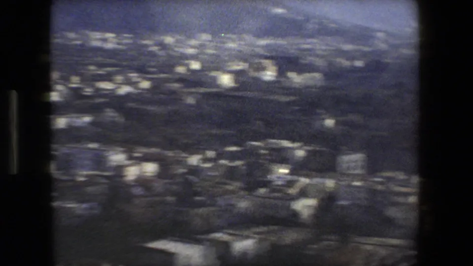 Blue-tinted 1970s 8mm frame showing a blurred Italian coastal townscape with scattered pale buildings and hillside contours.