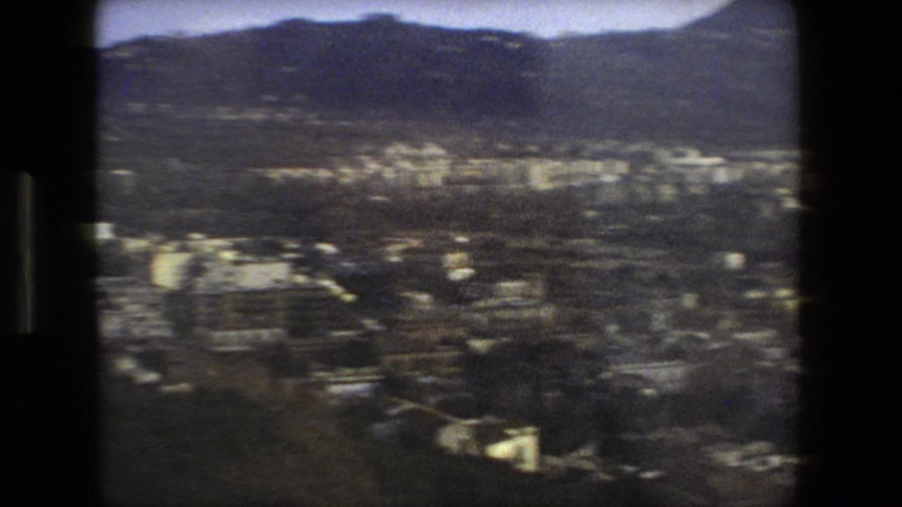 Grainy high-angle view over a dense city with dark hills in the background.