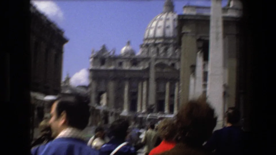 Soft handheld 1973 8mm frame from Rome showing tourists in the foreground and St. Peter's Basilica behind them, with the dome, obelisk, and surrounding architecture visible.