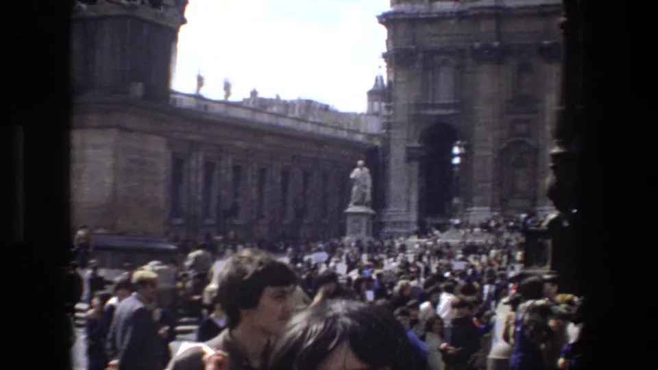 1973 Rome 8mm frame of a dense tourist crowd in St. Peter's Square, with a statue, colonnade, basilica architecture, and visitors in 1970s clothing visible.