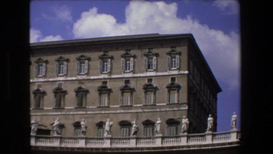 1973 8mm visitor view of a Vatican facade with repeated shuttered windows, colonnade statues, and blue sky visible behind the stone architecture.