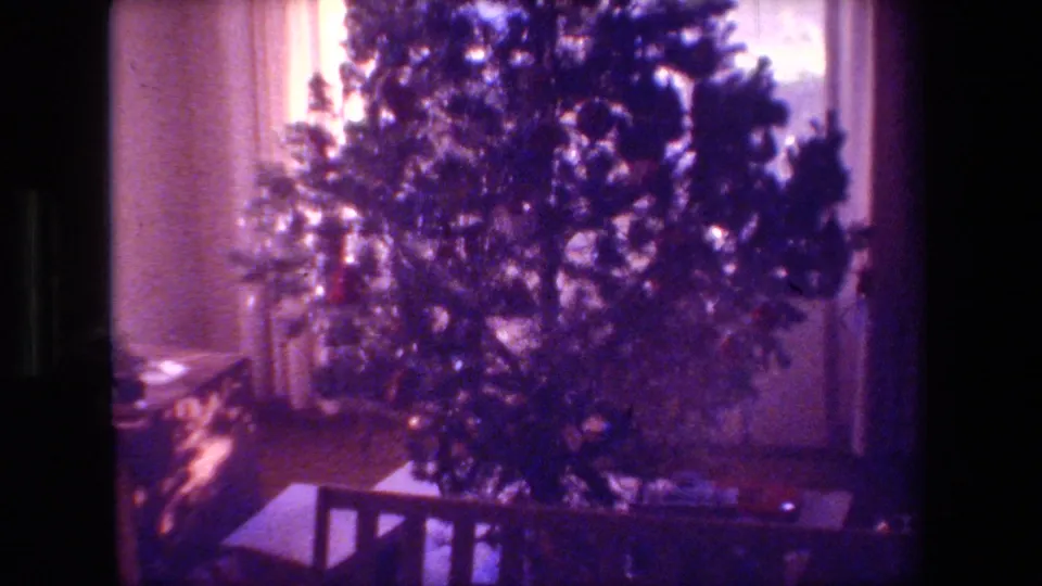 A decorated tree fills the living-room frame, with wrapped gifts near the base and a wooden bench or railing in the foreground.