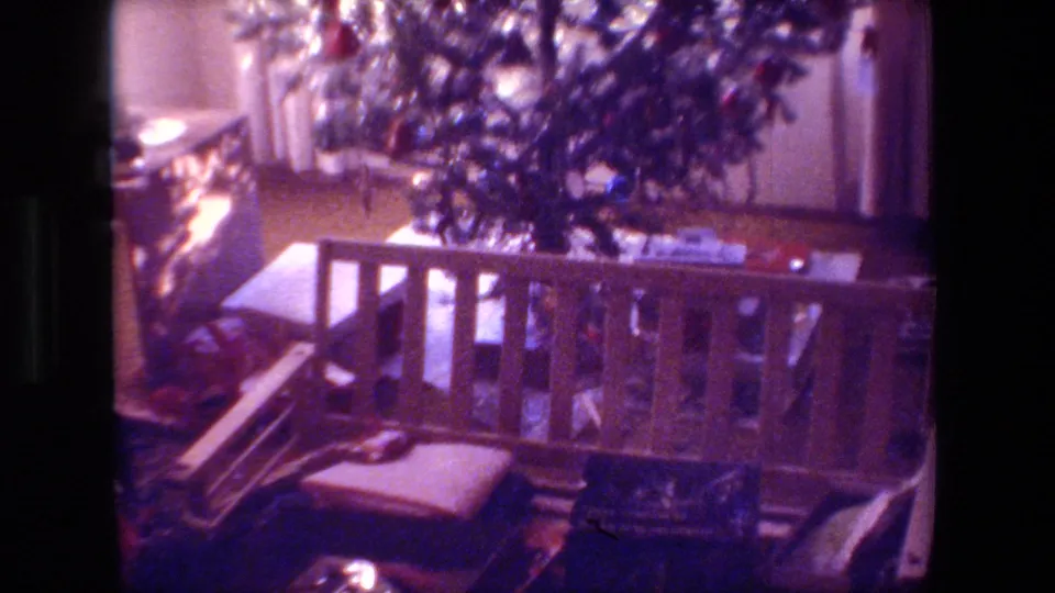 1981 Hollywood home-movie frame showing a dim private living room with a decorated Christmas tree, wooden bench or railing, cushion, and scattered household items.