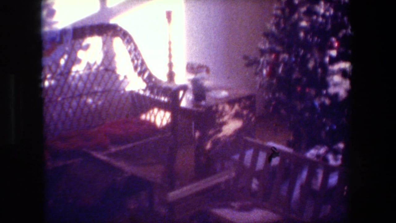 1981 Hollywood home-movie frame with a wicker chair by a bright window, a wooden bench or railing, and a decorated Christmas tree partly visible in a private room.