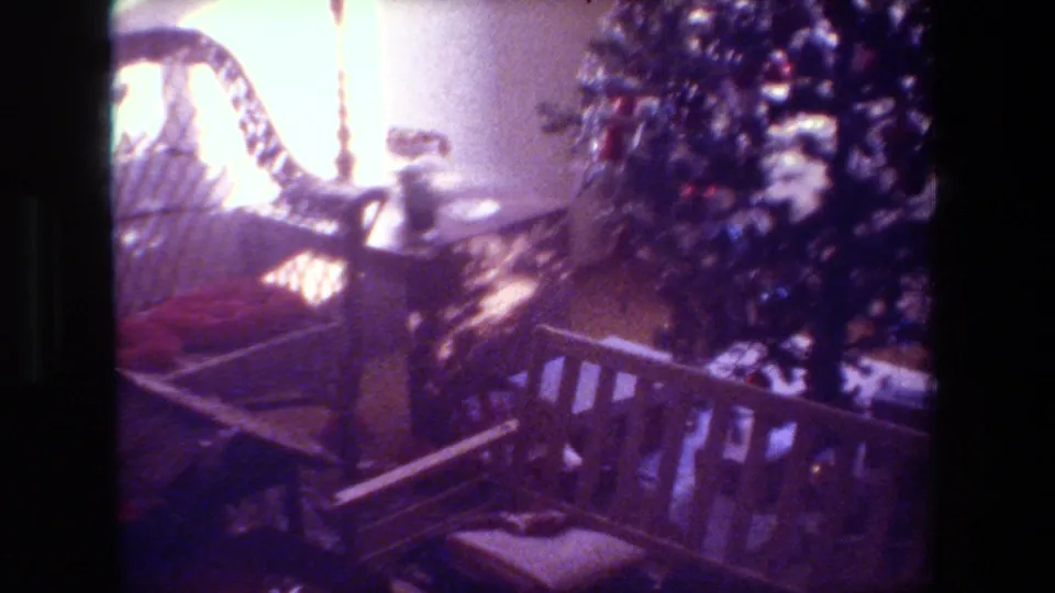 1981 Hollywood home-movie frame showing a wicker chair, decorated Christmas tree, wooden bench or railing, cushion, and scattered boxes under soft interior light.