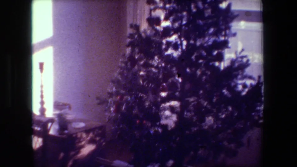1981 Hollywood home-movie frame centered on a decorated Christmas tree beside bright window light, with a candlestick and room furnishings partly visible.