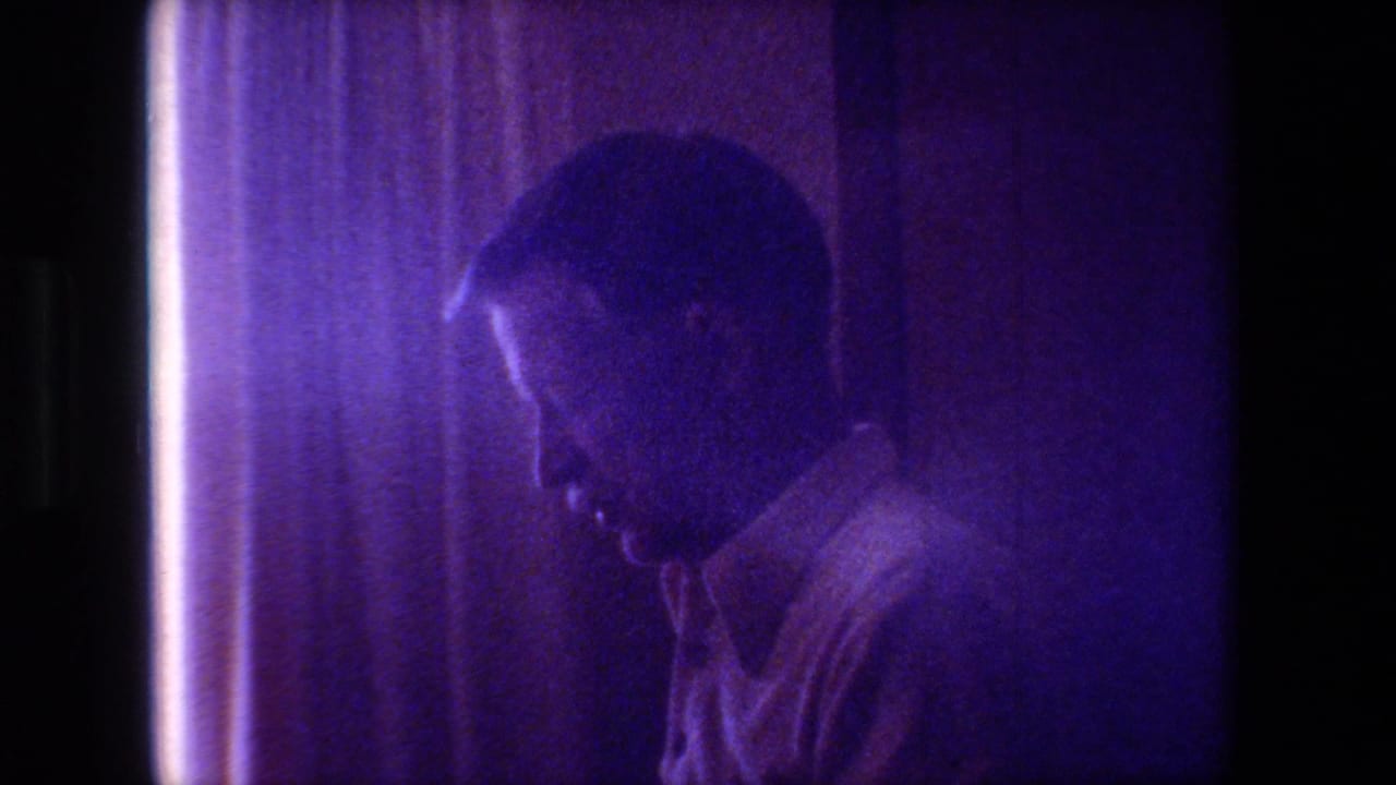 Adult man in profile near curtains in a dimly lit room.