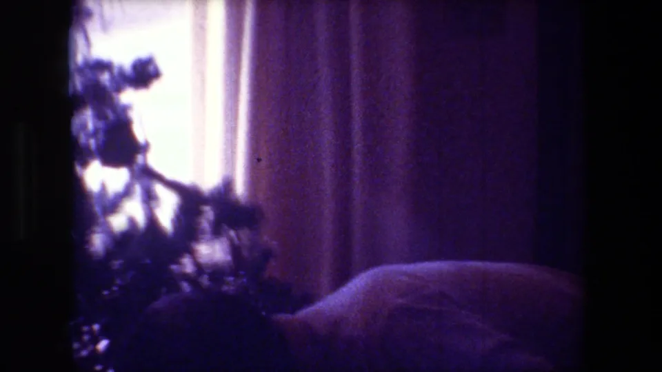 Soft, handheld 8mm framing shows blurred Christmas tree branches, curtains, and a low sofa or cushion inside the private holiday interior.