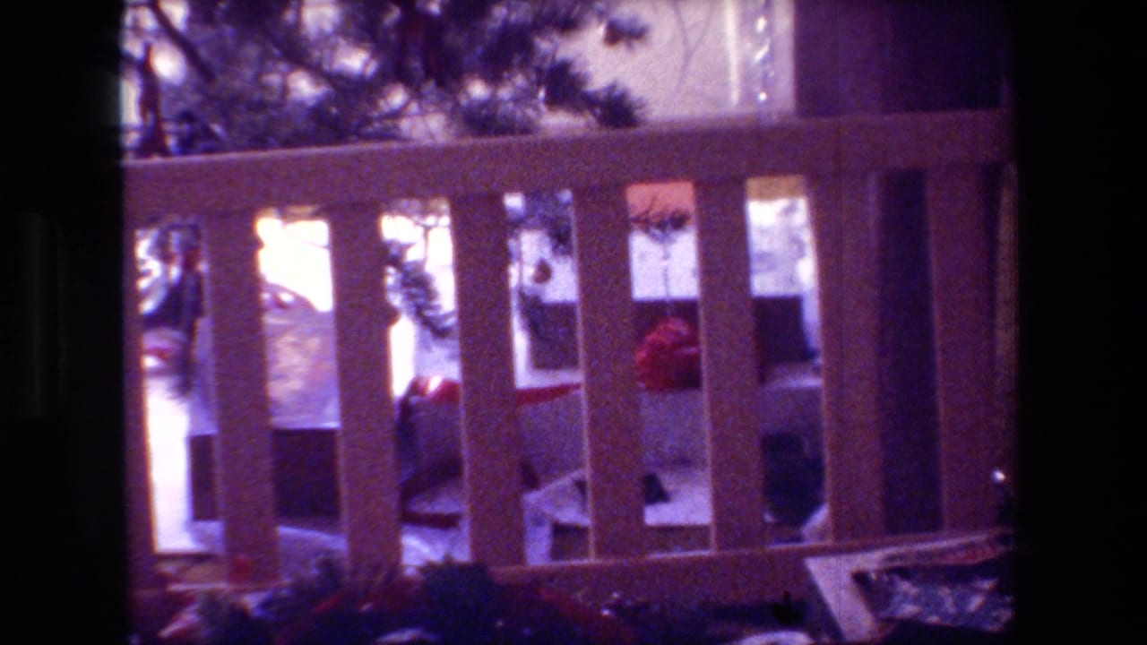 In this 1981 Hollywood, California home-movie frame, a small Christmas tree is seen near bright window light and a wooden rail inside a shadowed room.