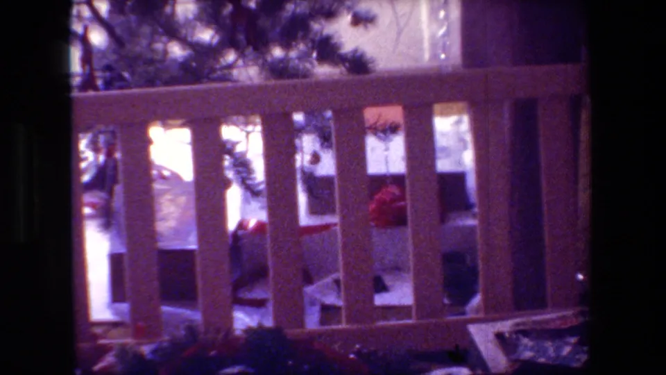 In this 1981 Hollywood, California home-movie frame, a small Christmas tree is seen near bright window light and a wooden rail inside a shadowed room.