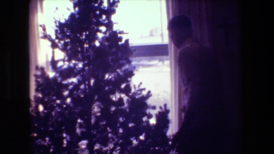 In this 1981 Hollywood, California home-movie frame, an adult man stands in shadow beside a small Christmas tree near a curtained window.