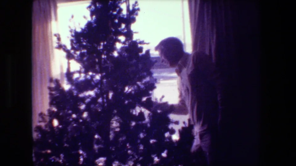 In this 1981 Hollywood, California home-movie frame, an adult man leans toward the small Christmas tree as if inspecting its branches beside the bright window.