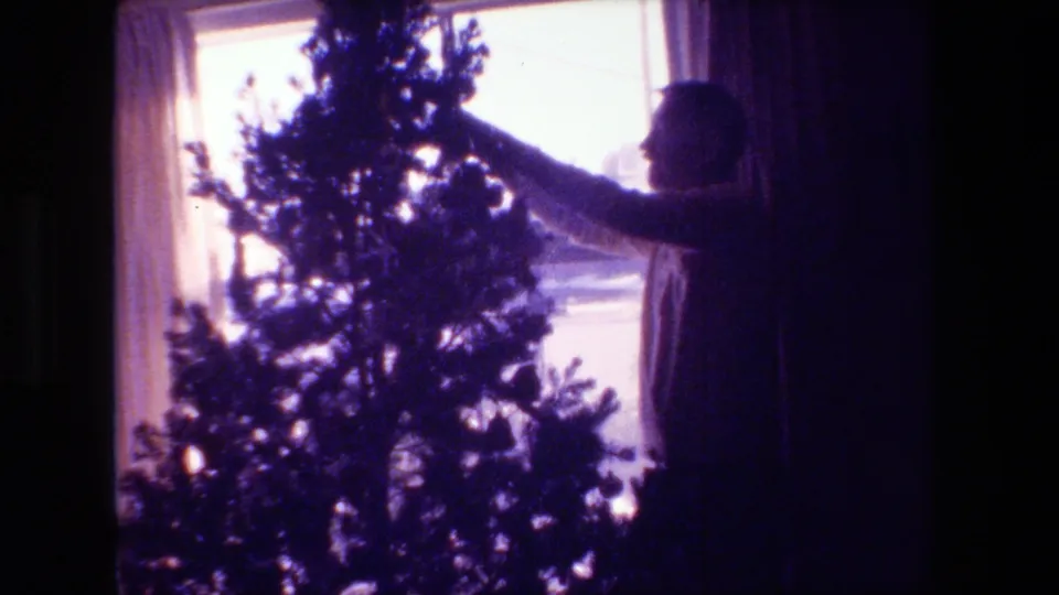 In 1981 Hollywood, California home-movie footage, a silhouetted adult man reaches up to decorate a Christmas tree positioned in front of a bright curtained window.