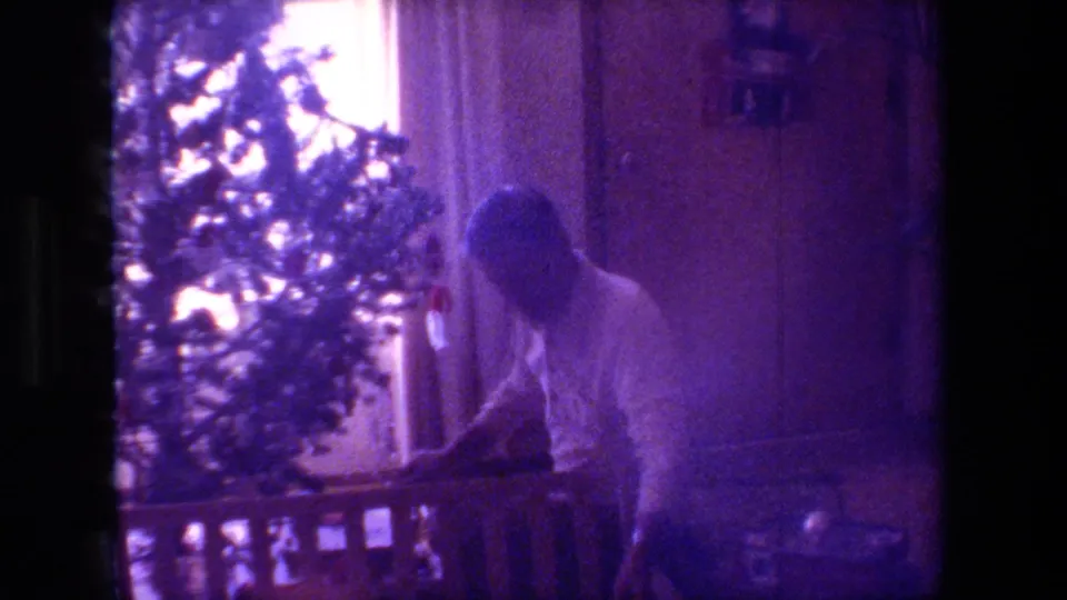 Inside the home, the man bends near the base of the Christmas tree and a low railing or furniture edge, with window light and wall paneling visible behind him.