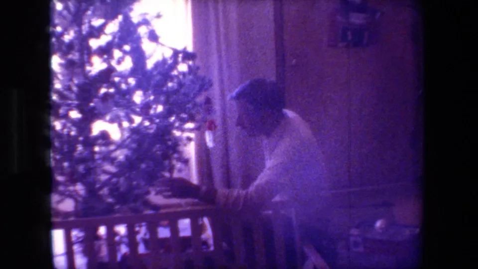 The man crouches or bends beside the indoor Christmas tree, with the bright window, curtains, wall paneling, and low railing or furniture edge framing the domestic holiday scene.