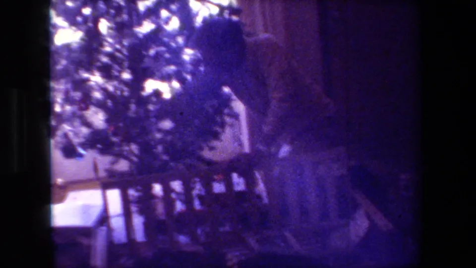 Adult man leans toward the Christmas tree in the dim living room, with the faded purple 8mm image showing the window and household furnishings around him.